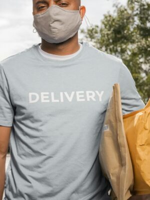 African American delivery man wearing a mask, holding parcels outside on a sunny day.