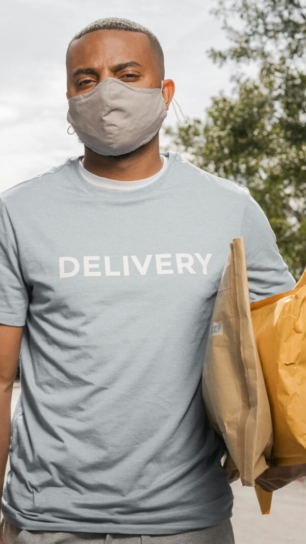 African American delivery man wearing a mask, holding parcels outside on a sunny day.