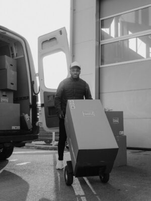 Monochrome image of a delivery man with boxes on a trolley near a van.